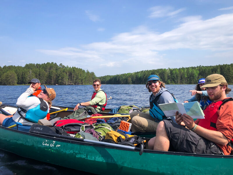 The image shows a group of people in a canoe on a lake. They are wearing life jackets and appear to be enjoying a sunny day. Some are looking at a map, suggesting they are navigating. The background features a forest and a blue sky.